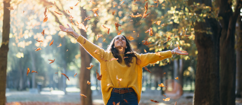 Casual joyful woman having fun throwing leaves in autumn at city park.