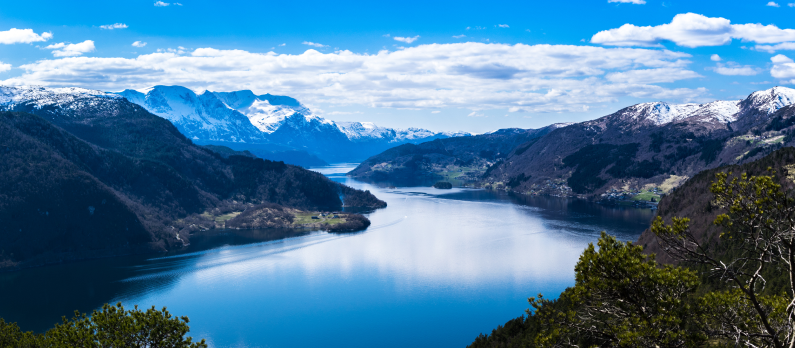 Oversiktsbilde av Sunnfjord med bl&aring;tt vann, gr&oslash;nne tr&aelig;r og fjellkjede i bakgrunnen