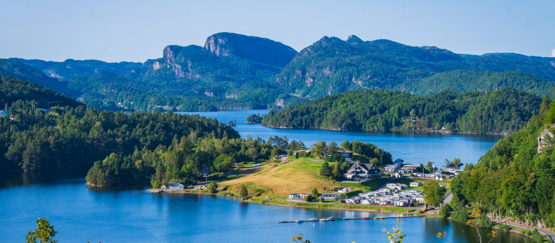 Oversiktsbilde over innsj&oslash;en Selura i Flekkefjord med bl&aring;tt vann omringet av bebyggelse, frodig skog og fjell i bakgrunnen